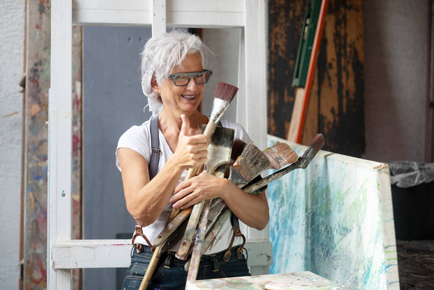 older gray haired mature happy artist woman with glasses and big paintbrushes gives thumbs up, copy space - Fotoğraf, Görsel