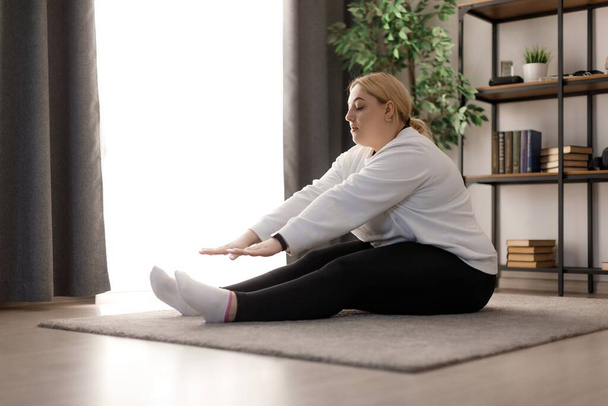 Приваблива молода жінка в спортивному одязі робить подовжені вправи, сидячи вдома на м "якому килимі. Білявка - білявка витрачає вільний час на тренування, щоб схуднути. - Фото, зображення
