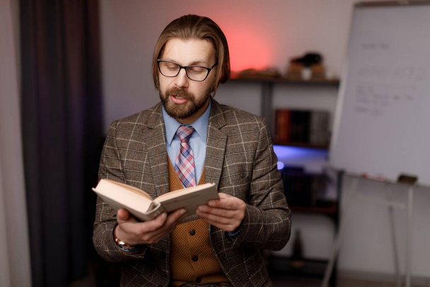 Uomo barbuto caucasico con gli occhiali che legge libro ad alta voce alla telecamera a casa studio. Concentrato blogger maschile in abbigliamento formale facendo live streaming con seguaci nei social network. - Foto, immagini