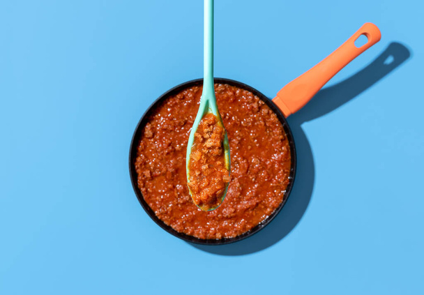 Above view with homemade bolognese sauce in a spoon minimalist on a blue table. Classic italian bolognese sauce stewed in a iron cast pot - Foto, Imagen