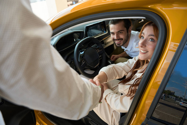 Salesperson shaking hands with joyful female client seated in car with her pleased companion - Foto, Imagen