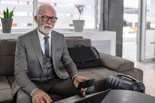 Portrait d'entrepreneur souriant avec téléphone intelligent et ordinateur portable. Heureux professionnel senior porte des lunettes et costume formel. Il est assis sur le canapé à table dans le bureau. - Photo, image