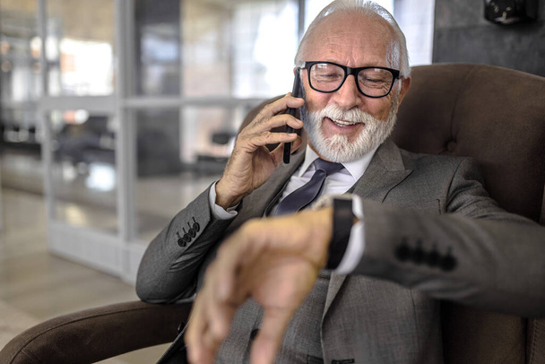 Lächelnder Unternehmer, der die Zeit auf der Armbanduhr kontrolliert. Ein älterer Geschäftsmann mit Brille spricht auf dem Smartphone. Er wartet an einer U-Bahn oder einem Bahnhof. - Foto, Bild