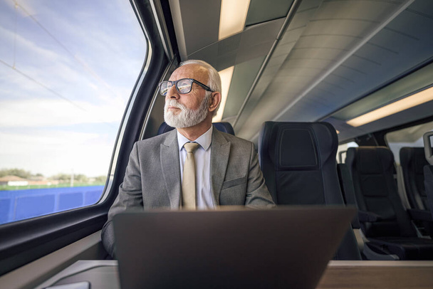Senior thoughtful businessman with laptop looking through window. Male professional is working while traveling on passenger train. He is wearing eyeglasses and formal suit. - Foto, Imagem