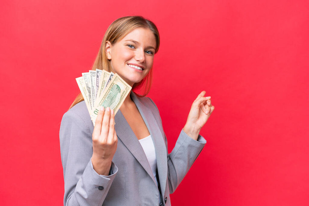jong bedrijf Kaukasische vrouw houden geld geïsoleerd op rode achtergrond wijzend terug - Foto, afbeelding