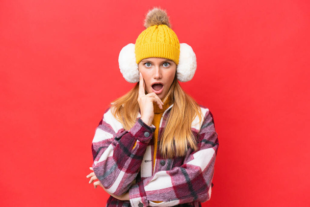 Молодая красивая женщина в зимних муфтах на красном фоне удивлена и шокирована, глядя направо - Фото, изображение