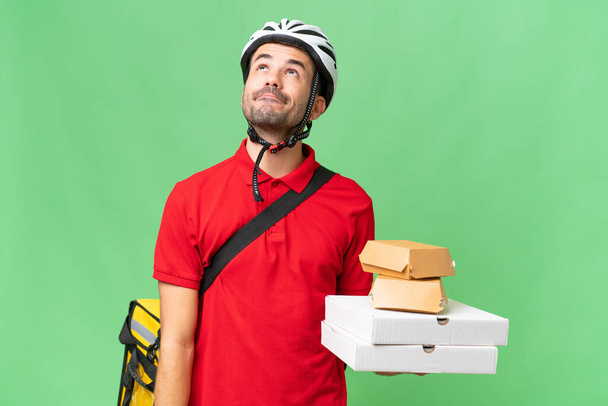 Young handsome caucasian man with thermal backpack and holding takeaway food over isolated background and looking up - Photo, Image