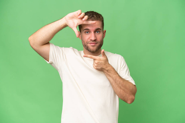 Jonge knappe blanke man geïsoleerd op groene chroma achtergrond gericht gezicht. Framing symbool - Foto, afbeelding