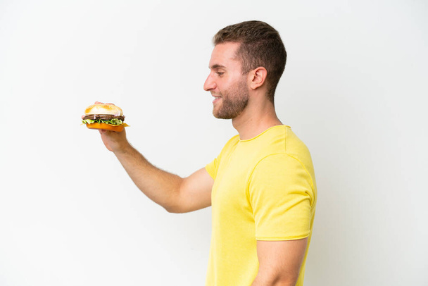 Young caucasian man holding a burger isolated on white background with happy expression - Fotó, kép