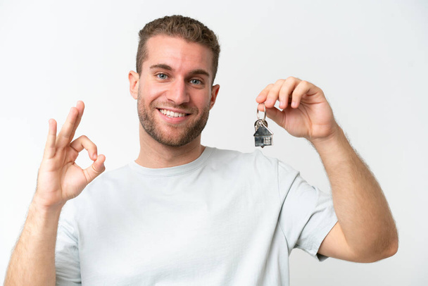 Young caucasian man holding home keys isolated on white background smiling and showing victory sign - Foto, imagen