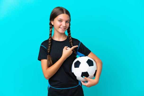 Pieni valkoihoinen tyttö eristetty sinisellä pohjalla jalkapallo pallo ja osoittaa sivusuunnassa - Valokuva, kuva