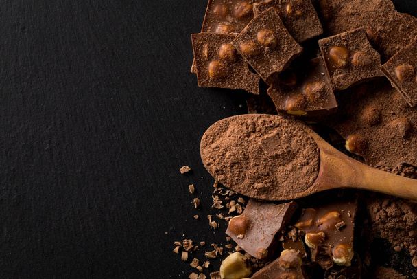 Piezas de nueces de chocolate rotas y cacao en polvo en cuchara sobre fondo de piedra oscura - Foto, imagen