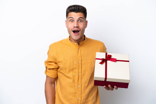 Young caucasian man holding a gift isolated on white background with surprise and shocked facial expression - Foto, Imagem