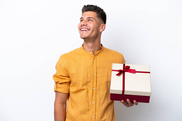 Jovem caucasiano segurando um presente isolado no fundo branco olhando para cima enquanto sorri - Foto, Imagem