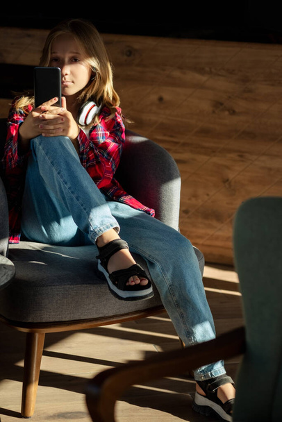 Teini-ikäinen tyttö rentoutua istua tuolilla kahvilassa matkapuhelimen tekstiviestien jutteleminen ystävän kanssa, onnellinen nuori nainen sohvalla pidä älypuhelin ostoksia verkossa tai tarkista mobiilisovellus tai sosiaalinen media - Valokuva, kuva