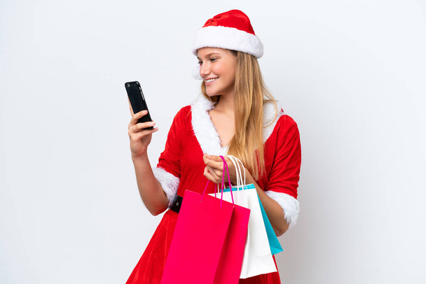 Young caucasian woman dressed as mama noel isolated on white background holding shopping bags and writing a message with her cell phone to a friend - Foto, afbeelding