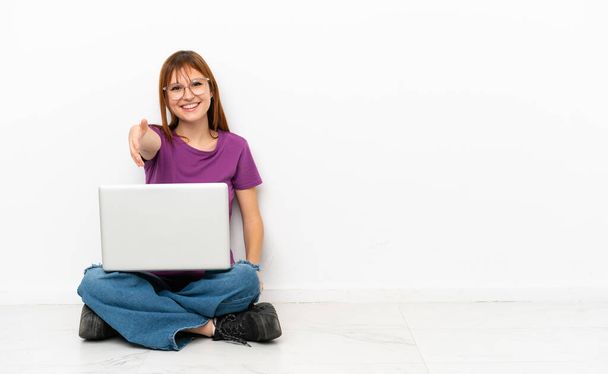 rothaariges Mädchen mit Laptop, das auf dem Boden sitzt und Hände schüttelt, um ein gutes Geschäft abzuschließen - Foto, Bild