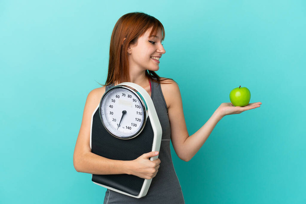 Redhead girl isolated on blue background holding a weighing machine while looking an apple - Photo, Image
