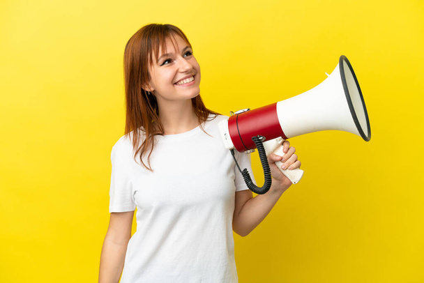 Redhead girl isolated on yellow background holding a megaphone and looking up while smiling - Foto, imagen