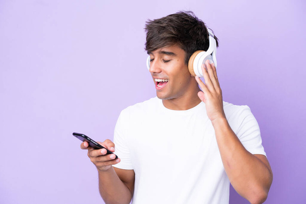 Fiatal kaukázusi férfi elszigetelt lila háttér hallgat zenét egy mobil és énekel - Fotó, kép