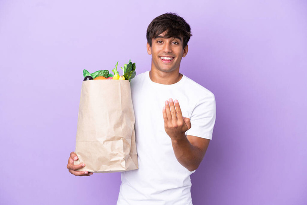 Молодой человек, держащий сумку с продуктами, изолированную на фиолетовом фоне, приглашает прийти с рукой. Рад, что ты пришла. - Фото, изображение