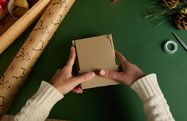 Top view female hands holding a cardboard box, packing gifts for Christmas or New Year event, on a green background with wrapping decorative meterials. Diy presents. Boxing Day. Handwork art craft - Photo, Image