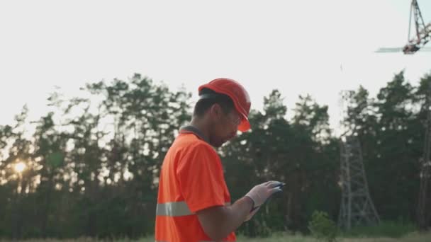 Mies yllään kypärä ja oranssi työvaatteet toimii tabletti voimalaitoksessa kentän lähellä korkea jännite tornit. Sähköinsinööri tarkistaa voimajohdon tabletin sähköantureiden tietojen avulla.  - Materiaali, video