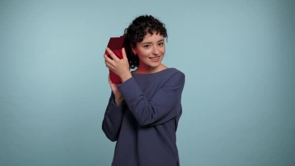 Schöne lächelnde lockige Frau schüttelt umhüllte rote Schachtel in der Nähe des Ohres interessiert, was drinnen ist. Positive Frau trägt blauen Pullover mit Geschenk in den Händen auf isoliertem hellblauem Hintergrund - Filmmaterial, Video