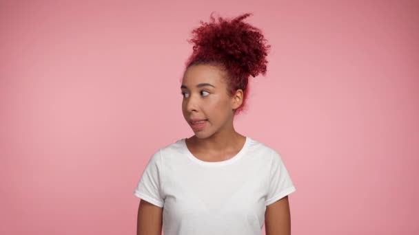 肖像の赤毛アフリカ系アメリカ人の女性の周りの唇の上に人差し指を置き、沈黙のジェスチャーを秘密を伝えます。コピースペースのあるピンクの背景に立つ白いTシャツを着たアフリカの女性 - 映像、動画