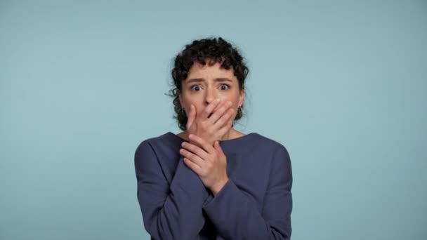 Portrait beautiful shocked curly woman covers mouth with palm looking camera. Excited scared female wearing blue sweater standing on isolated light blue background with copy space. Embarrassment - Footage, Video