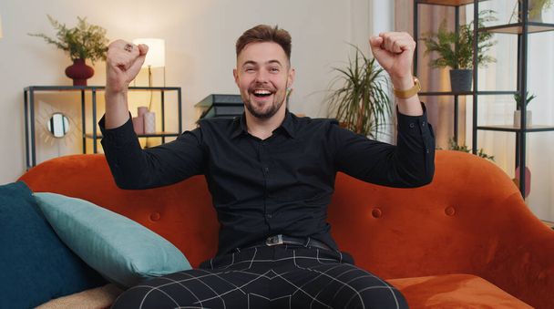 Happy caucasian man shouting, celebrating success, winning, goal achievement, good news, raising fists in gesture I did it, lottery luck at home apartment. Young guy indoors in living room on couch - Foto, Imagem