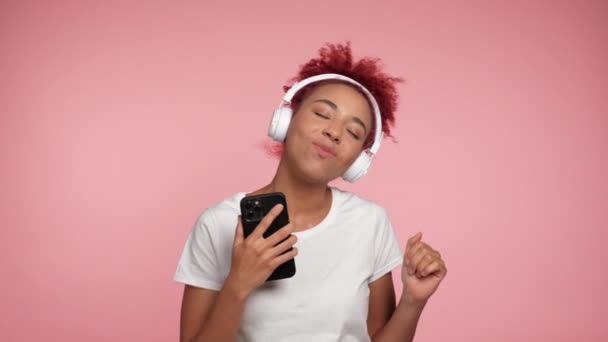 Close up mosolygós afro-amerikai vörös hajú göndör nő tartja okostelefon a kezében zenét hallgat fejhallgató, táncok. Portré boldog pozitív szép nő elszigetelt rózsaszín háttér másolási hely - Felvétel, videó
