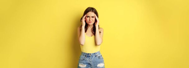 Stylish modern woman rubbing temples, having headache, migraine, frowning and grimacing from pain in head, standing against yellow background. - Foto, imagen