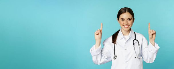 Портрет уверенной молодой женщины врач, медицинский работник в пальто, указывая пальцами вверх и улыбаясь, показывая рекламу лекарств, промо клиники, токуазный фон. - Фото, изображение