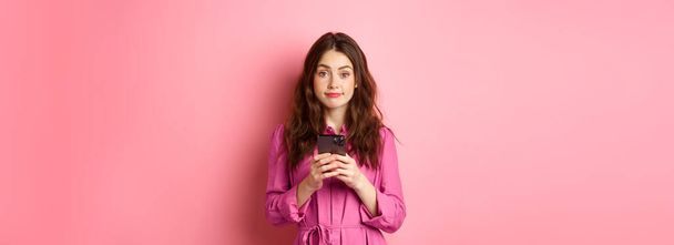 Cute young woman using mobile phone app, holding smartphone and smiling at camera, standing in dress against pink background. Copy space - Photo, Image