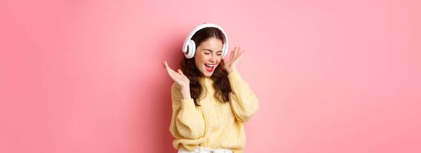 Chica feliz cantando canción favorita, escuchando música en auriculares inalámbricos, sonriendo y bailando, de pie sobre fondo rosa. - Foto, Imagen