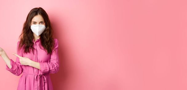 Ковід-19, концепція пандемії та способу життя. Стильна сучасна дівчина в респіраторі, спрямовуючи пальці вліво, показуючи копійку в сторону, стоячи на рожевому фоні
. - Фото, зображення