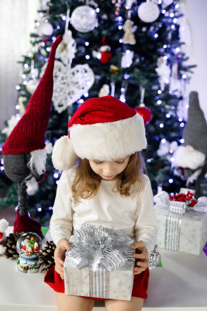 Uma menina em um chapéu de Papai Noel vermelho está sentado sob a árvore de Natal e brincando com uma caixa de presente. Ambiente de Natal e Ano Novo. Conceito de uma infância feliz. - Foto, Imagem