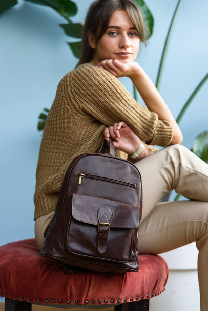 beautiful slender girl in beige pants and olive sweater posing while sitting with a small brown leather backpack. Indoor photo - Foto, Imagen