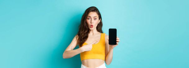 Intrigued girl asking about smartphone app, pointing at mobile screen and look curious at camera, standing against blue background. Copy space - 写真・画像
