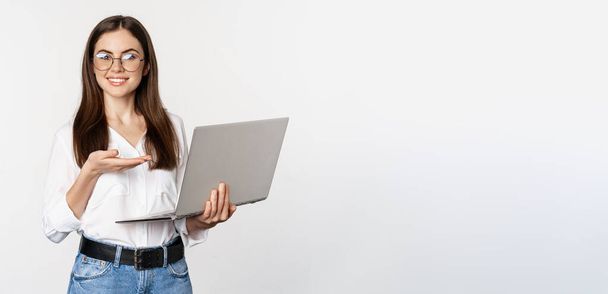 Retrato de mujer en gafas sosteniendo portátil, apuntando a la pantalla, mostrando su trabajo en el ordenador, de pie sobre fondo blanco. - Foto, imagen