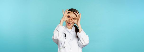 Funny therapist, girl doctor showing okay, binoculars zero gesture on eyes and smiling, fooling around and having fun, standing over blue background. - Foto, Bild