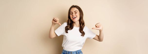 Self-assured young happy woman pointing fingers at herself and dancing, self-promoting, being confident, standing over beige background. - Foto, afbeelding