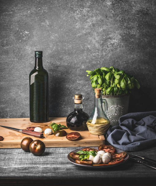 Still life with Italian caprese salad served on rustic table with potted basil kitchen herbs and bottle of olive oil on rustic table with ingredients - Valokuva, kuva