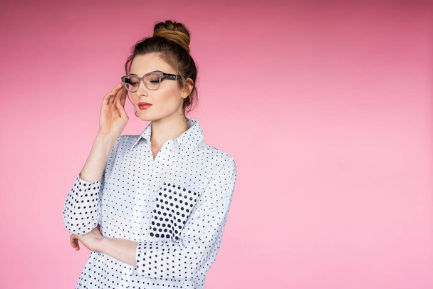 Linda modelo feminina vestindo camisa branca e óculos posando em fundo rosa em estúdio. Olhando para o lado e segurando uma mão perto do rosto. - Foto, Imagem