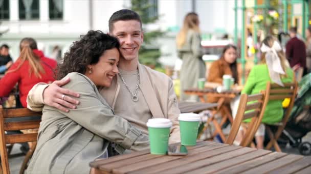 Una pareja feliz al aire libre cerca de un café. Abrazándose, sonriendo, tomando café. Ambiente otoñal. Movimiento lento - Imágenes, Vídeo