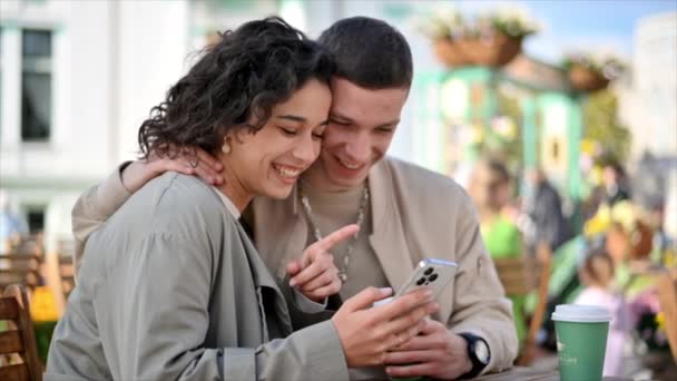 Una pareja feliz al aire libre cerca de un café. El hombre está abrazando a su mujer, mirando por teléfono, sonriendo, hablando, tomando café. Ambiente otoñal. Movimiento lento - Imágenes, Vídeo