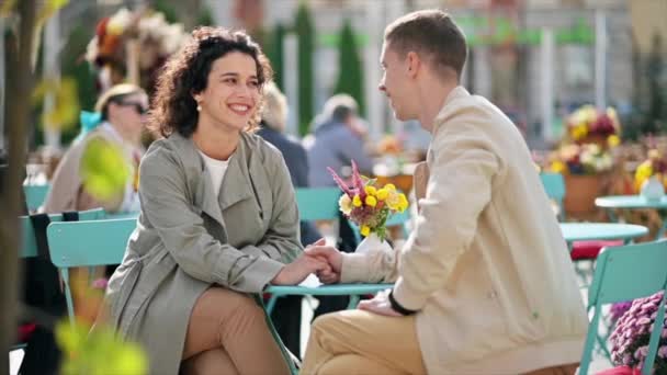 Una pareja romántica al aire libre cerca de un café. Tomados de la mano, sonriendo, hablando y mirándose, flores en la mesa. Ambiente otoñal. Movimiento lento - Imágenes, Vídeo