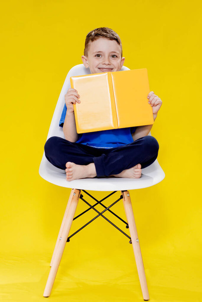 Little cheerful blond green-eyed boy 5-6 years old in a stylish blue T-shirt holding book and reading on yellow wall background, children's studio portrait. - Photo, Image