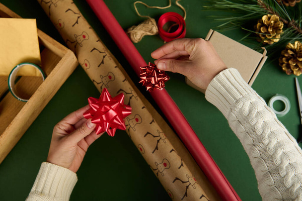 トップビューの女性の手は、クリスマスや新年の贈り物を梱包するための材料と緑の表面に、鹿のパターンと赤の色の包装紙に結び弓を適用します。ボクシングの日。手芸だ。死ぬんだ - 写真・画像